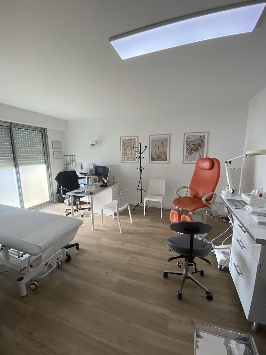 Sa salle: le fauteuil de podologie orange, la table d'examen, le bureau blanc et trois cadres de fleurs séchées au mur.